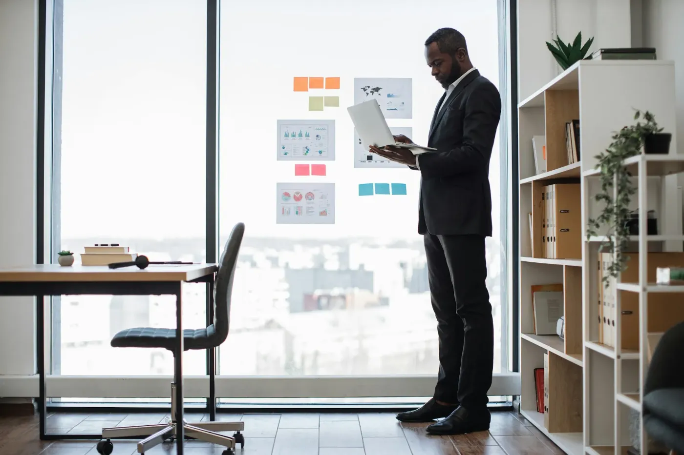 Man with a laptop standing at a floor-to-ceiling office window