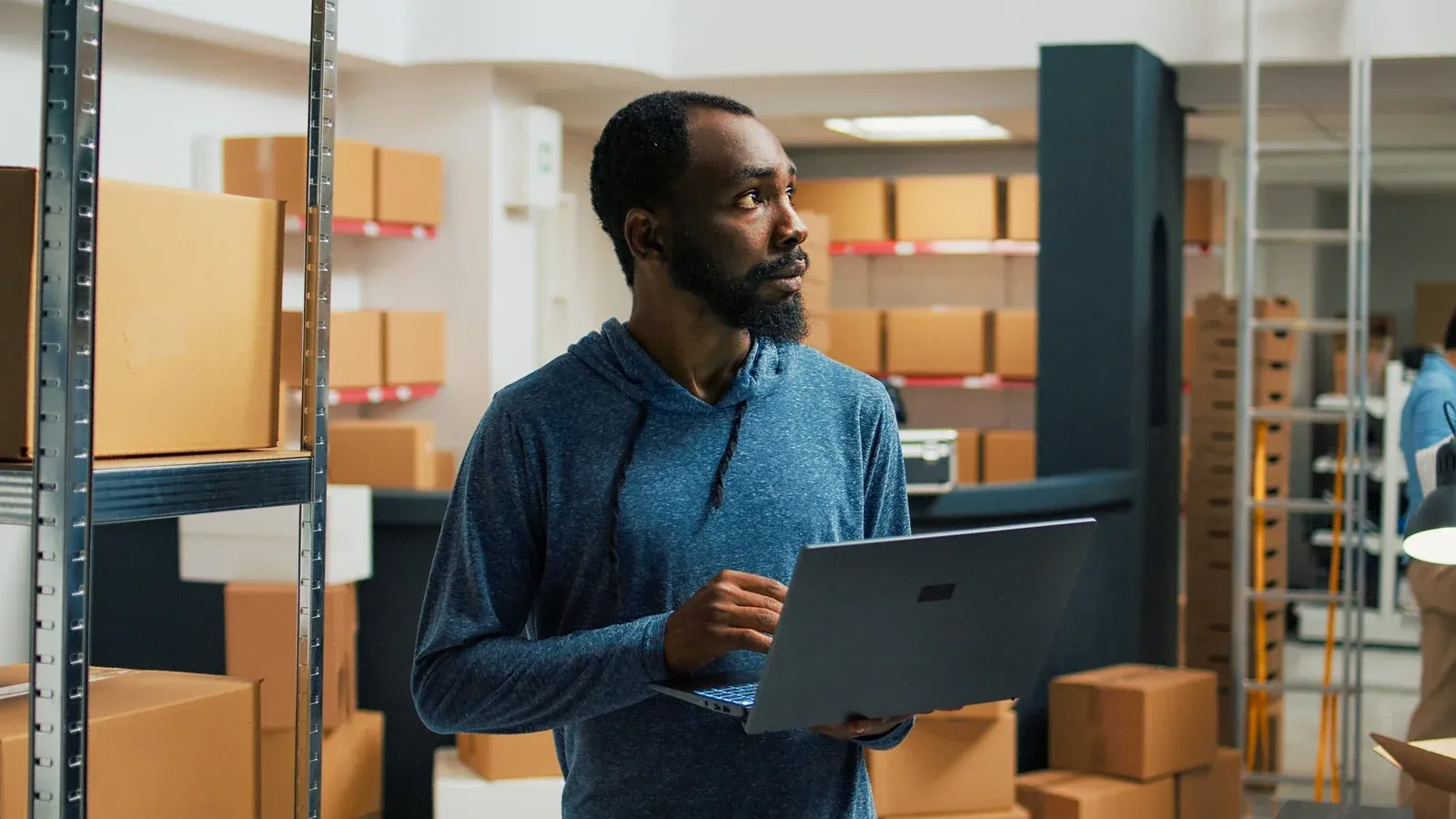 Warehouse worker checking stock on a laptop between shelves of boxes