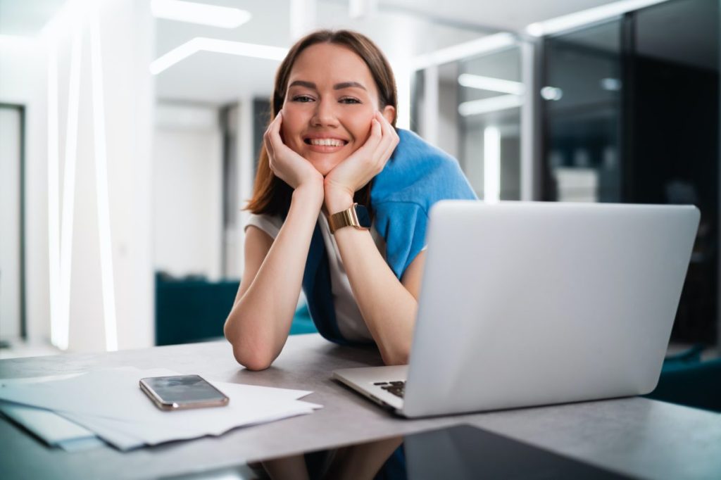 Happy smiling young woman sitting at desk with open laptop looking at camera. Successful smiling