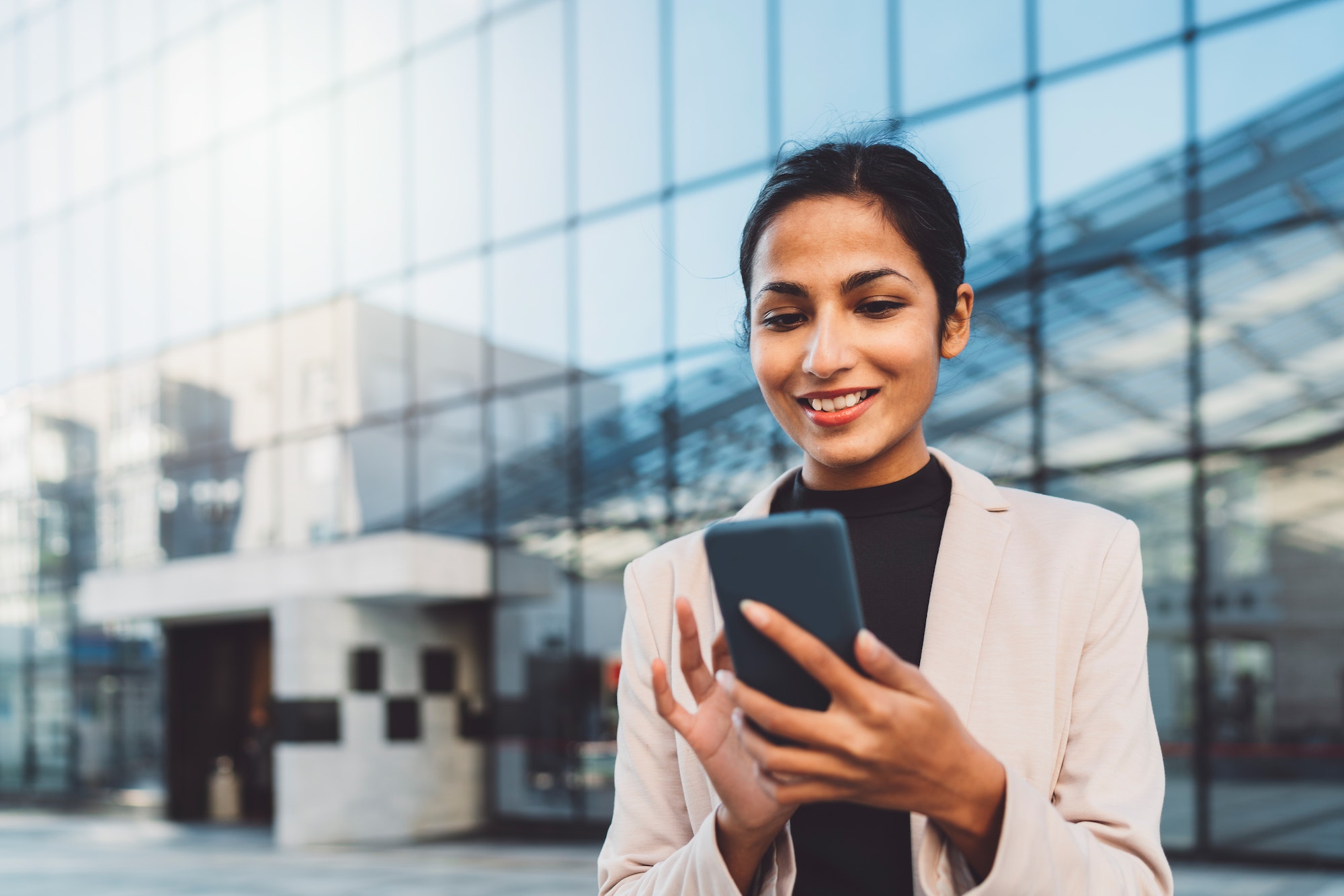 Smiling young business woman writing an email on her phone while standing outside on the sun during