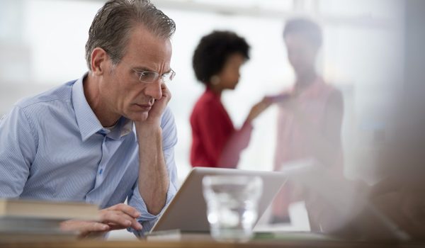 A man seated at a computer laptop leaning on one arm, and two women in the background.