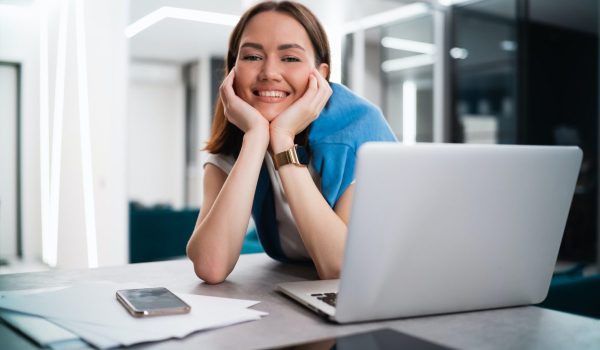 Happy smiling young woman sitting at desk with open laptop looking at camera. Successful smiling