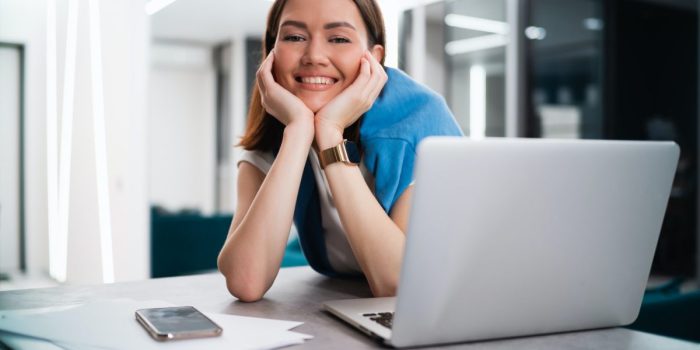 Happy smiling young woman sitting at desk with open laptop looking at camera. Successful smiling