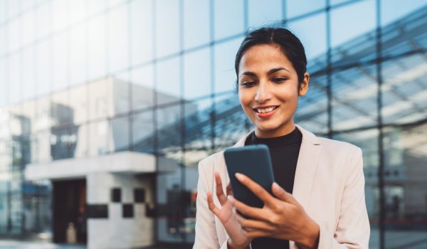Smiling young business woman writing an email on her phone while standing outside on the sun during