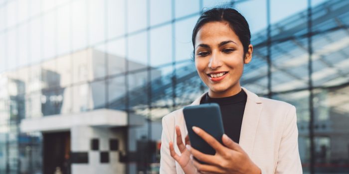 Smiling young business woman writing an email on her phone while standing outside on the sun during